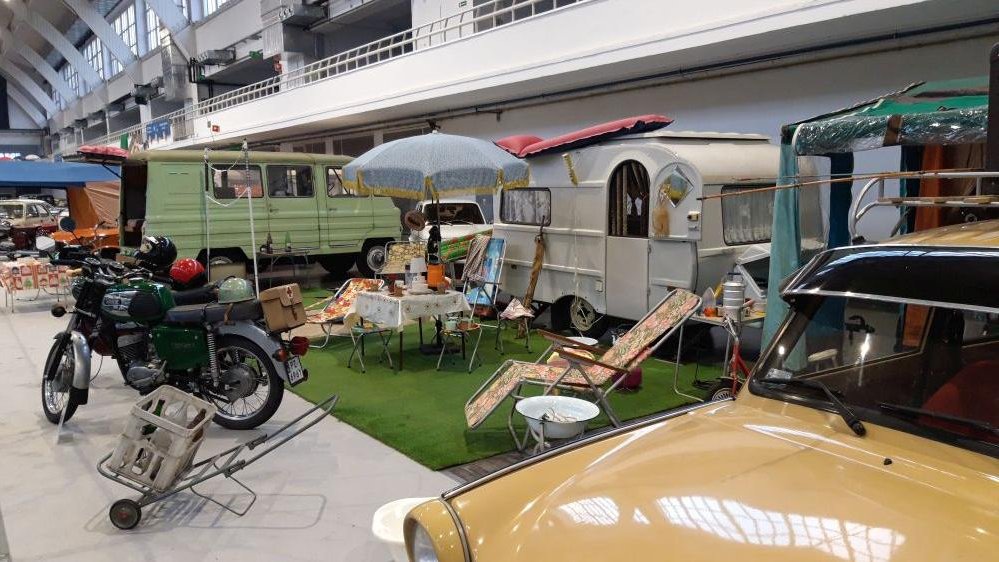 The photo from the exhibition: an old motorbike and an old caravan. Between them a table with oilcloth on it and a few old fashioned deck chairs.