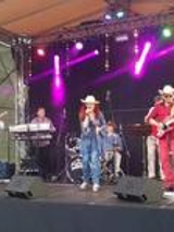Picture of a country group - singing woman and two men playing the guitars on a stage. In the background a man playing drums and a man playing keyboard.