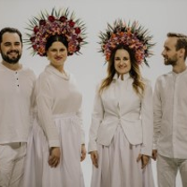 The photo shows four smiling musicians from the Dagadana band. Everyone in stylized white costume, with colorful wreaths of flowers on their heads, white wall as a background