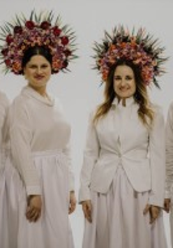 The photo shows four smiling musicians from the Dagadana band. Everyone in stylized white costume, with colorful wreaths of flowers on their heads, white wall as a background