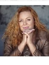 The photo shows a smiling, golden-haired artist looking straight into the camera. She rests her chin on her folded hands.