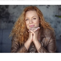 The photo shows a smiling, golden-haired artist looking straight into the camera. She rests her chin on her folded hands.