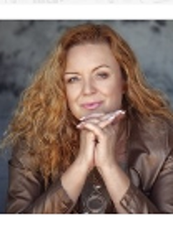 The photo shows a smiling, golden-haired artist looking straight into the camera. She rests her chin on her folded hands.