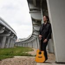 A man leaning against bridge pillar, holding a guitar