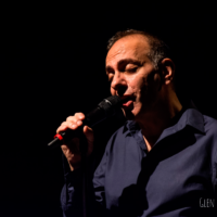 Photograph of the artist - a man in a shirt holding a microphone and singing. Black background.