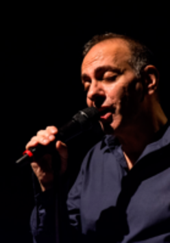 Photograph of the artist - a man in a shirt holding a microphone and singing. Black background.