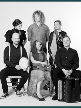 The black and white photograph shows the six members of the Okra team in two rows. In the first one, a woman and two men are sitting on chairs. Behind them are two more ladies and one gentleman. Some of them hold musical instruments.