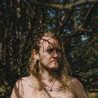 Photo presents face and shoulders of a long-haired man. Tree branches in the background