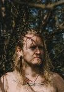 Photo presents face and shoulders of a long-haired man. Tree branches in the background