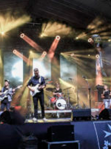 A colourful photo of a band on a stage: two men playing the guitars, a man playing drums, two singing women