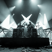 The black and white photo taken during the concert, from under the stage. In its central part there are two musicians - one plays the banjo and the other plays the bagpipes.
