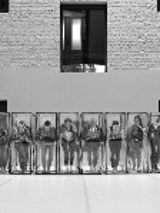 Black and white photo of the dancers in a row, who are standing in transparent boxes.