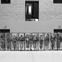Black and white photo of the dancers in a row, who are standing in transparent boxes.
