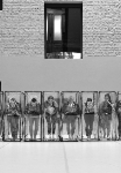 Black and white photo of the dancers in a row, who are standing in transparent boxes.