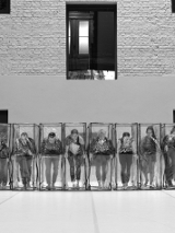 Black and white photo of the dancers in a row, who are standing in transparent boxes.