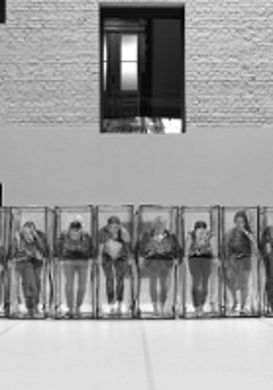 Black and white photo of the dancers in a row, who are standing in transparent boxes.