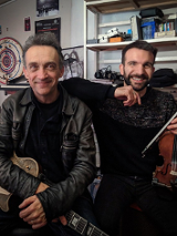 Photo of the artists: two men looking into the camera, smiling and holding their musical instruments. Shelves with CDs and various things in the background