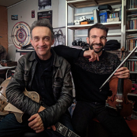 Photo of the artists: two men looking into the camera, smiling and holding their musical instruments. Shelves with CDs and various things in the background