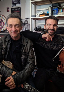 Photo of the artists: two men looking into the camera, smiling and holding their musical instruments. Shelves with CDs and various things in the background