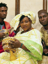 The photo shows the artists: one woman and two men. The woman is wearing a beautiful long dress in shades of light yellow and green, and a decorative turban on her head. She is holding a musical instrument in her hands. The other artists, dressed in folk costumes, are sitting next to her.