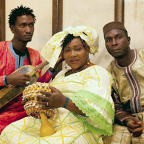 The photo shows the artists: one woman and two men. The woman is wearing a beautiful long dress in shades of light yellow and green, and a decorative turban on her head. She is holding a musical instrument in her hands. The other artists, dressed in folk costumes, are sitting next to her.