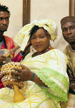 The photo shows the artists: one woman and two men. The woman is wearing a beautiful long dress in shades of light yellow and green, and a decorative turban on her head. She is holding a musical instrument in her hands. The other artists, dressed in folk costumes, are sitting next to her.