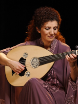Photo of a woman in a long dress. The artist sits and holds an oval stringed instrument in her hands