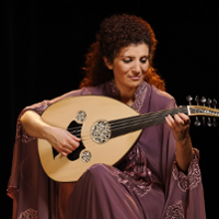 Photo of a woman in a long dress. The artist sits and holds an oval stringed instrument in her hands