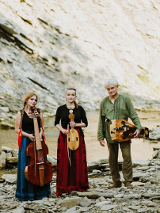 The photo shows three musicians, two women and a man, in the background there is a rocky hill. Musicians stand in front of the camera holding musical instruments