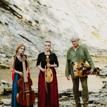 The photo shows three musicians, two women and a man, in the background there is a rocky hill. Musicians stand in front of the camera holding musical instruments