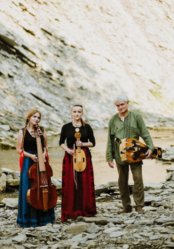 The photo shows three musicians, two women and a man, in the background there is a rocky hill. Musicians stand in front of the camera holding musical instruments