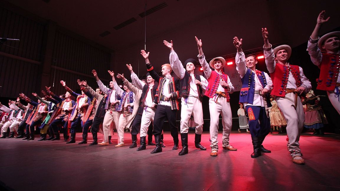 Photo of a folk group in traditional folk costumes performing on stage - dozen or so men standing in a row with their right hands up.