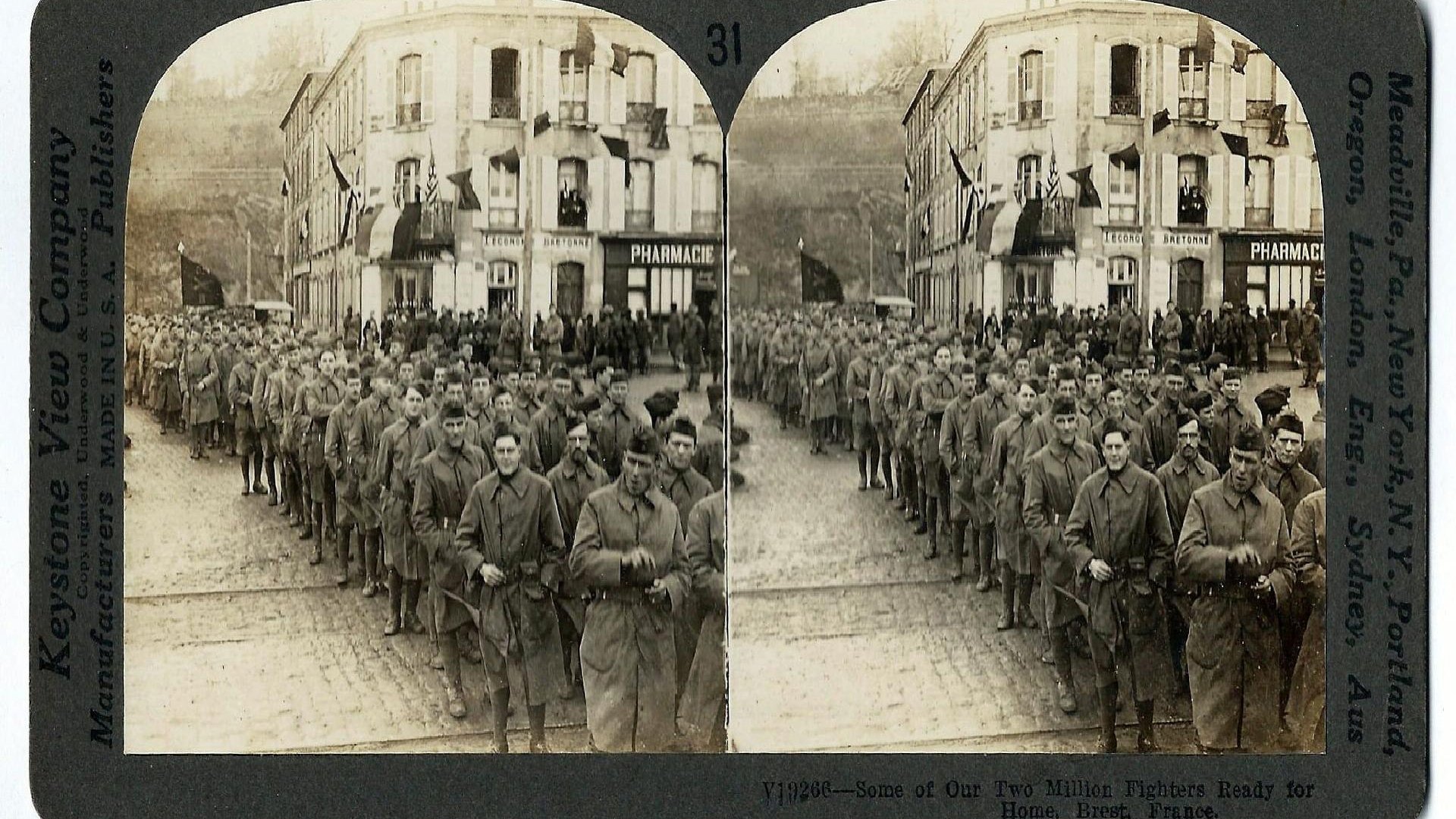 Photo in sepia colours of soldiers standing in rows on a cobbled street. A building in the background.