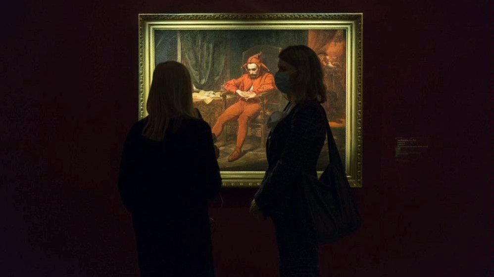 Photo of illuminated picture from the exhibition (the picture titled "Stańczyk" by Jan Matejko) on a black wall. Black outline of two people standing in front of the picture.
