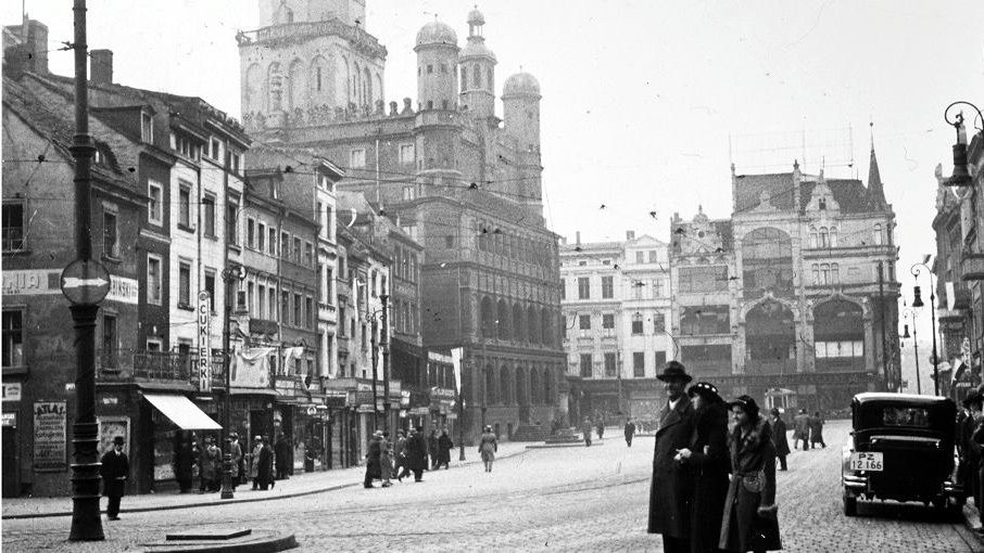Black and white picture of the Old Market in Poznań. People walking on the Old Market Square. The Poznań Town Hall and tenement houses in the background.