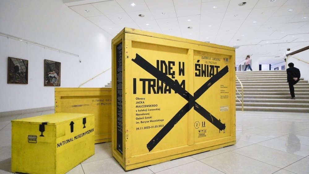 Photo from the exhibition: yellow shipping boxes with black inscriptions, stairs in the background on the right, two small paintings hanging on the left.