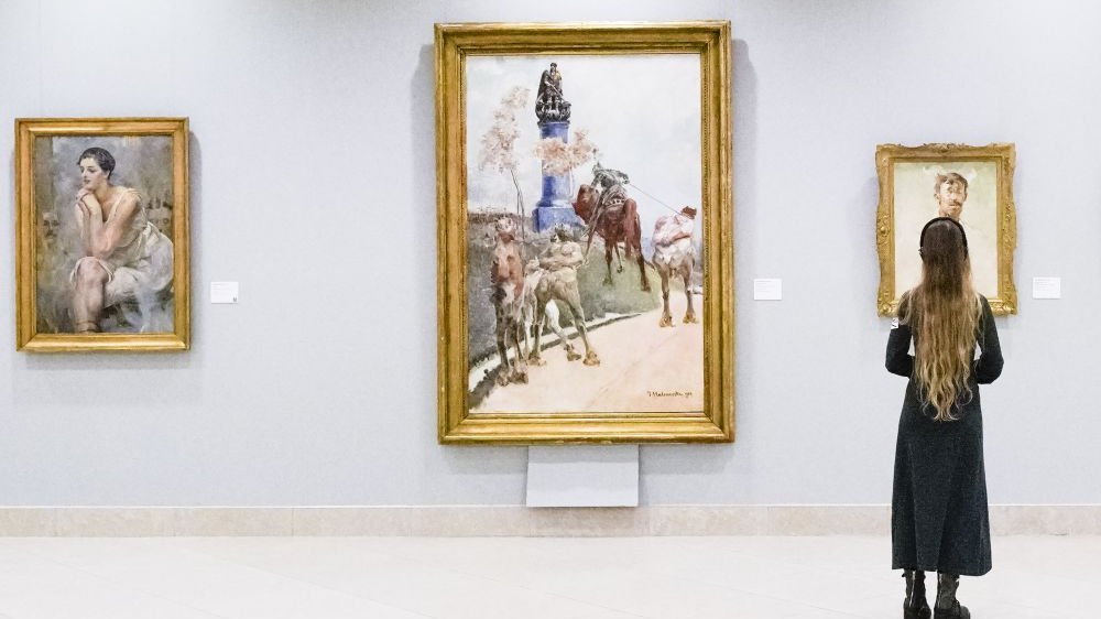 Photo of an exhibition hall with three paintings on the wall. In front of the paintings, the person who is looking at them.