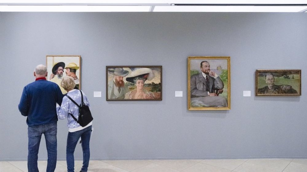 Photo of an exhibition hall with four paintings on the wall. In front of the paintings, the couple who is looking at them.
