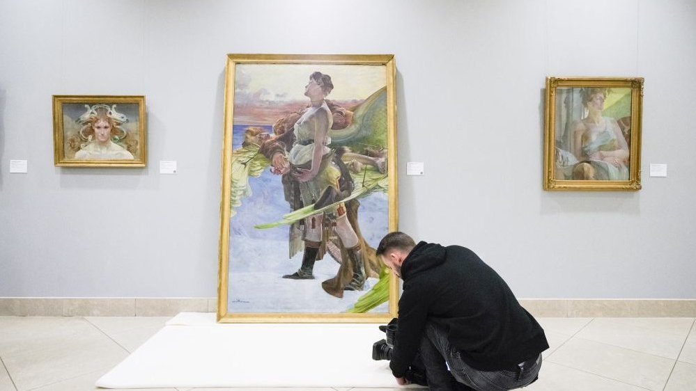 Photo of an exhibition hall with three paintings - two hanging and one big leaning against the wall. A man is taking a photo of a big picture.
