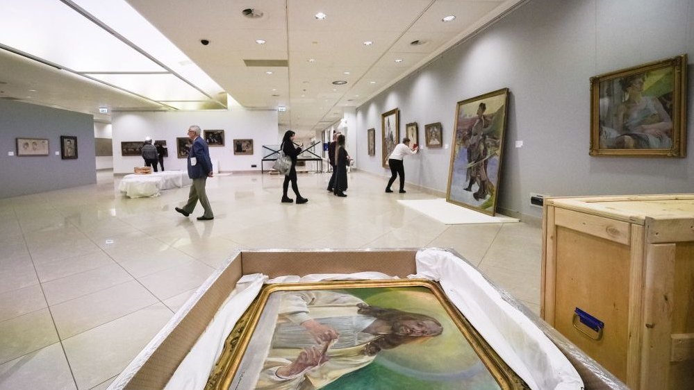 Photo of the exhibition hall. In the foreground there is a painting lying in a chest. In the background people watching the exhibition.