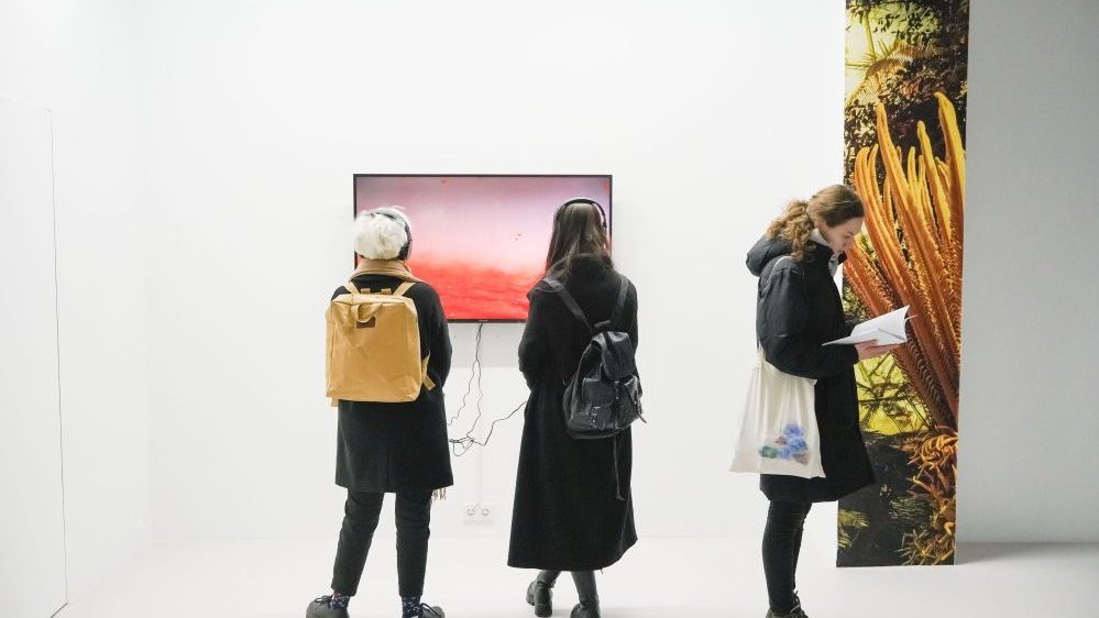 Exhibition hall: two people watching the picture displayed on a TV screen and one person reading a book.