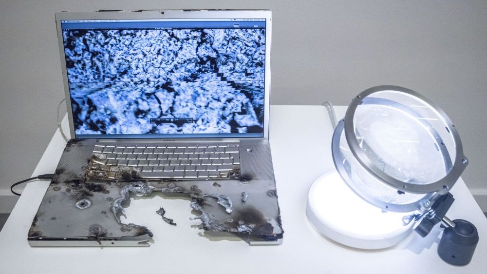 Photo of a partly burnt laptop standing on a white table; a kind of a lamp next to the laptop.