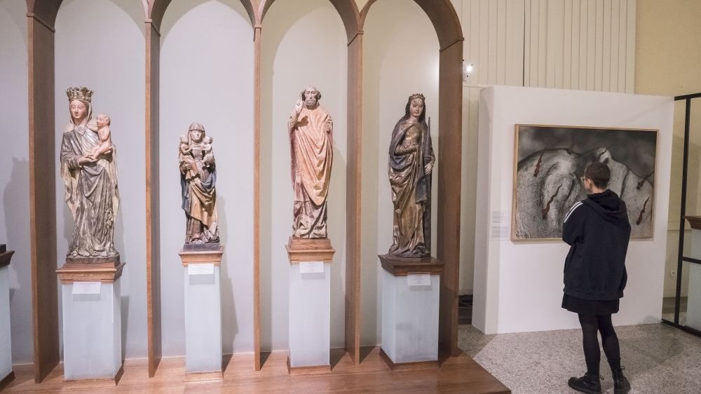 Four wooden figures of different sizes placed next to each other. Three of them depict images of Mary. Next to them there is a large graphic painting depicting a mutilated face.