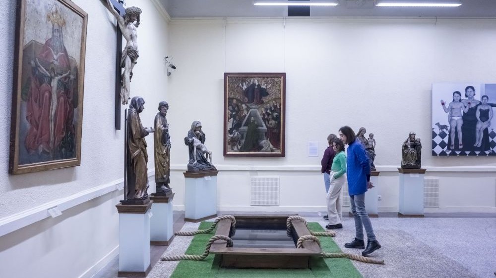 Viewers look at paintings and sculptures at the exhibition. One of the paintings is attached to the floor, one has to bend down to see it.