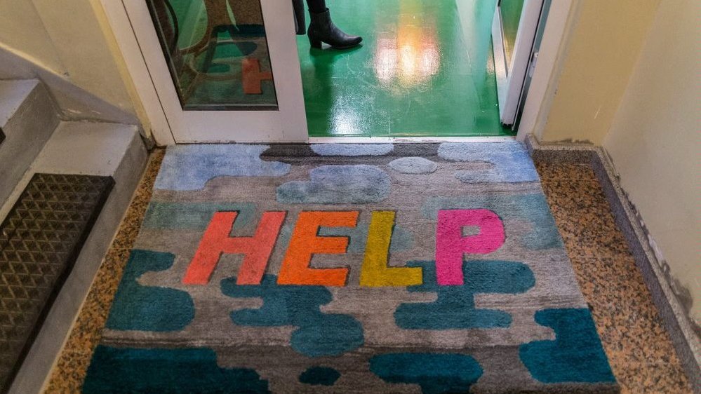 One of the artist's works - a colourful doormat with an inscription "Help" on it in front of the door.