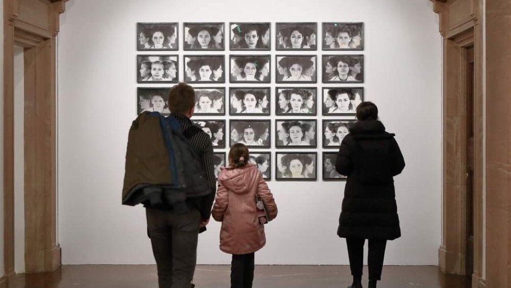 Photo of black and white exhibition works hanging on the wall and three people looking at them.