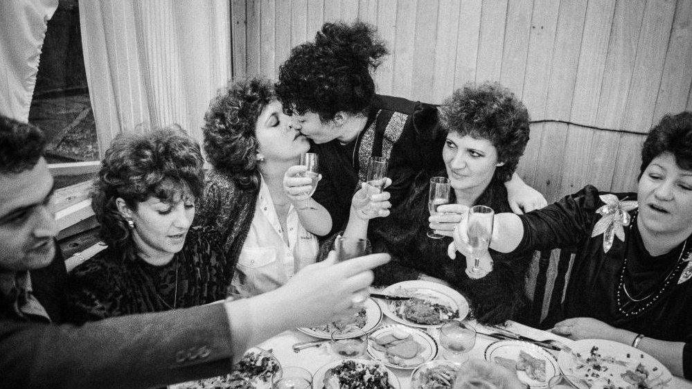 Black and white photo of a few women and a man sitting at the table with glasses in their hands. Two women are giving each other a kiss.