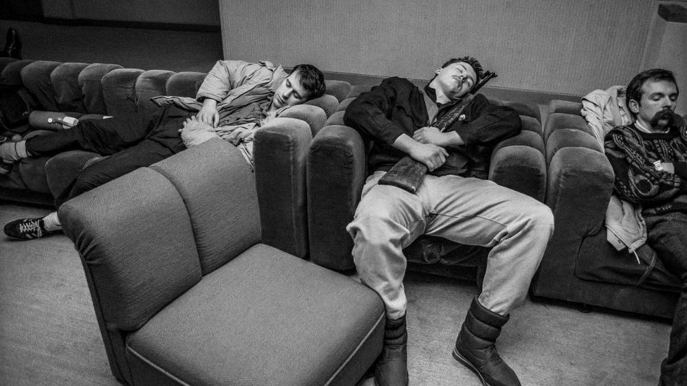 Black and white photo of a three men sleeping in the arm-chairs. One of them is holding a rifle.
