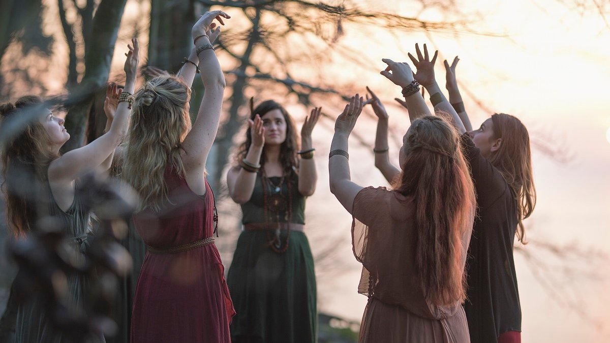 Photo of a band: seven women standing in a circle with their hands raised.