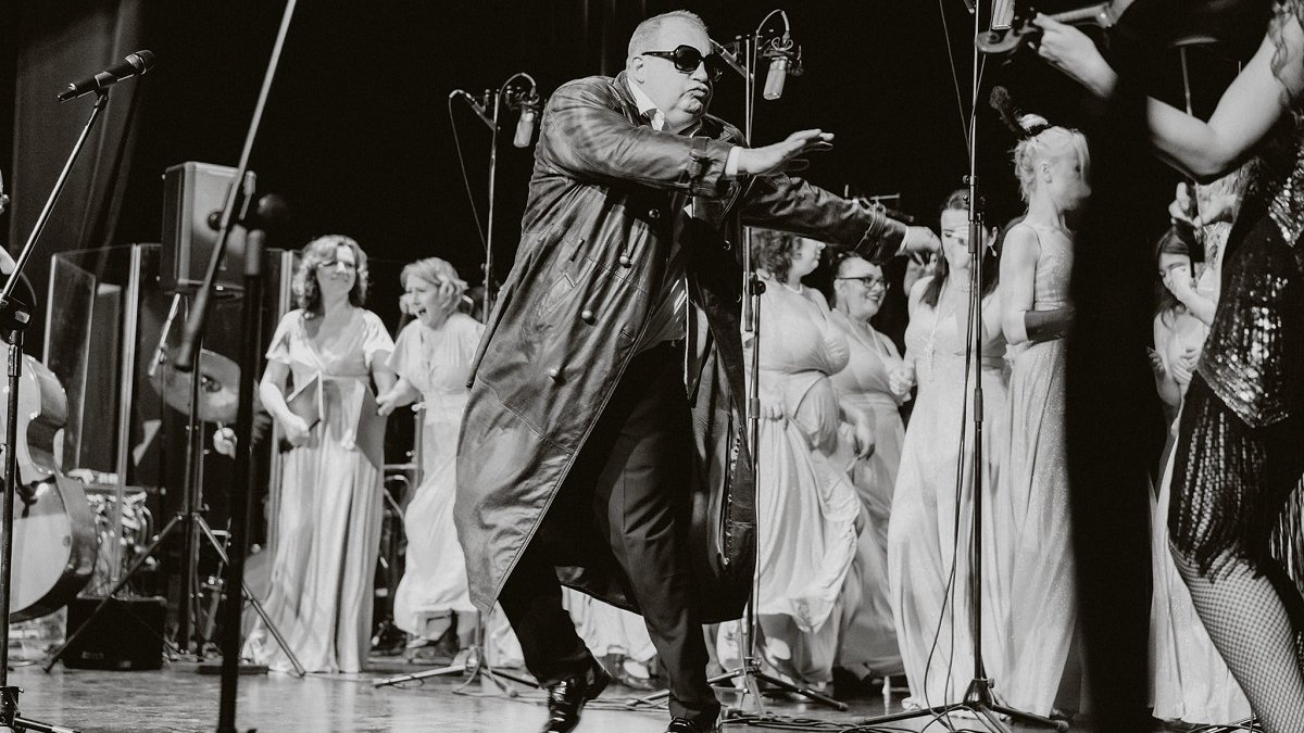 A black and white photo from a concert: a man in sunglasses and a black leather coat performing on stage, behind him women in long white dresses.